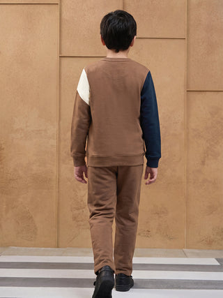 BOYS, standing and walking away from the camera, wearing a brown long-sleeve sweatshirt with color-block panels in white and navy blue, and matching brown sweatpants.