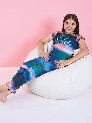 A young girl poses cross-legged on a beanbag, wearing a colorful tie-dye sleep set with star prints.
