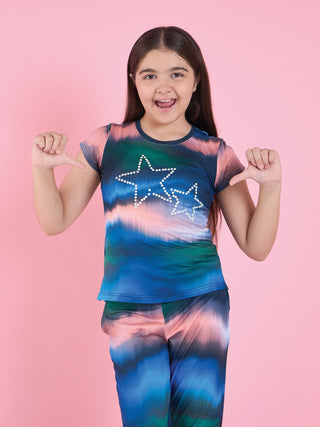 Girl standing front-facing against a pink backdrop pointing at herself, wearing a navy short-sleeve pyjama set with blue, teal and peach tie-dye print and pearl star embellishment.