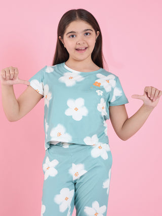 Girl standing against a pink backdrop pointing at herself, wearing a green short-sleeve pyjama set with white floral print and orange flower details.