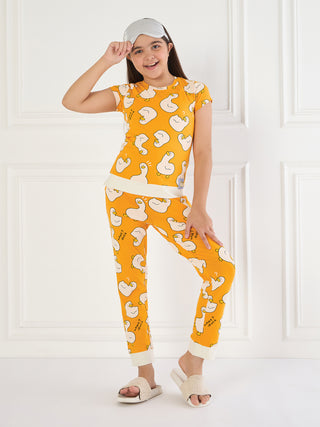 A young girl in an orange pajama set with a duck print, a white sleep mask, and white slippers stands in front of a white wall.