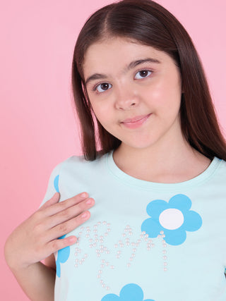 Girl posing in a close-up with one hand on her chest, wearing a crystal green short-sleeve sleepwear top with blue flower print and pearl embellishment.