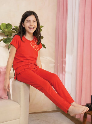 A young girl in a red pajama set with gold heart print, sitting on a couch.