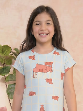 A young girl in a light blue, short-sleeved nightgown with a grid pattern and scattered teddy bear prints smiles at the camera.