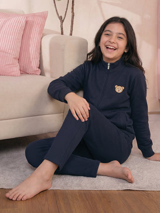 A young girl in a navy blue pajama set with a teddy bear print sits on the floor, smiling.