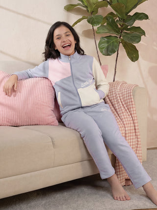 A young girl with dark hair sits on a couch wearing a color-blocked lavender, pink, and cream track suit.