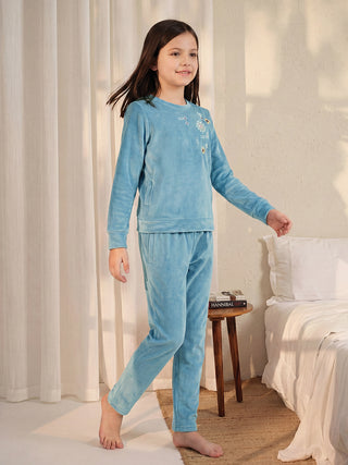 A young girl stands with her left leg forward, wearing light blue velour pajamas with floral embroidery.
