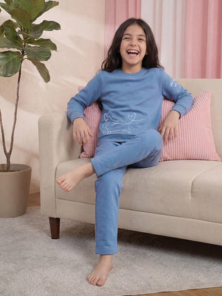 A young girl in a blue pajama set with a dog print smiles sitting on a couch.