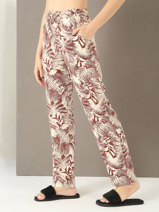 Women wearing relaxed fit, cream and brown palm-print pajama pants and black fuzzy slides, with one hand in her pocket.