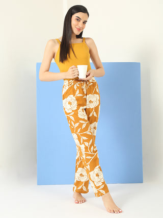 A woman stands holding a mug, wearing a mustard yellow tank top and yellow floral print pants.