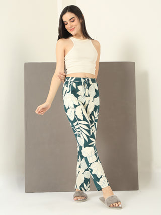 A woman in a cream ribbed tank top and green floral print lounge pants poses with her right arm crossed over her chest.