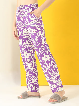 Women's purple pajama pants with a white floral print, shown from the waist down, with one hand in a pocket, standing on a white and yellow background, wearing gray furry slippers, similar to the previous image but with a different pajama print.
