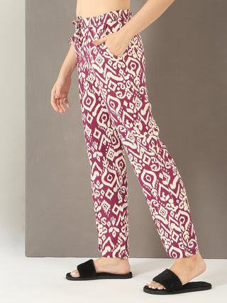 Women's burgundy and cream ikat print pajama pants, side view with hands in pockets and black fluffy slides.