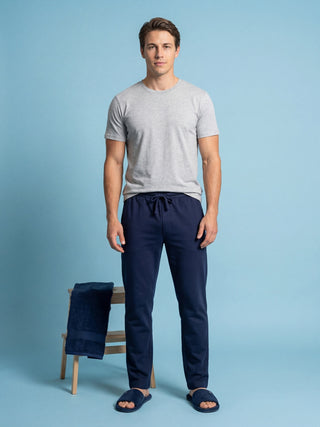 Men's grey t-shirt and navy blue loungewear pants, standing with a towel on a stool and slippers on his feet.