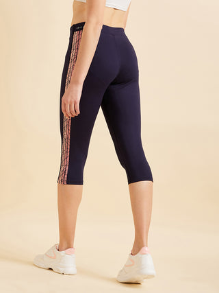 Women in a standing pose wearing navy blue capri leggings with a pink geometric side stripe.