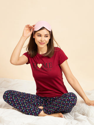 A woman sits cross-legged on a bed wearing a maroon t-shirt with a heart graphic and "HOME" text, and navy blue pajama pants with a colorful heart print, while holding a pink eye mask.