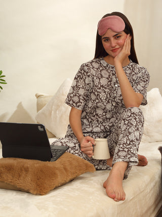 A woman in brown floral print sleepwear with a pink eye mask on, sitting on a couch with a laptop and mug.