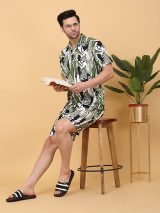 A man sits on a white chair wearing a pink and blue abstract print pajama set, holding a green mug.