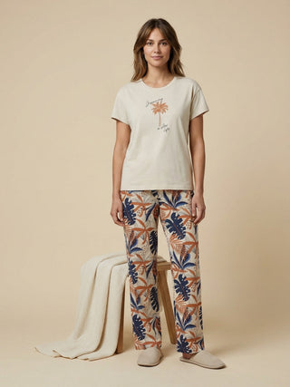 Women in a relaxed pose, wearing a cream-colored t-shirt with a palm tree graphic and "dreaming a slow life" text, paired with patterned cream, navy blue, and rust-colored pajama pants.