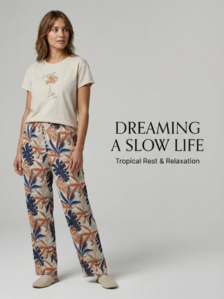 A woman poses in cream loungewear with a palm tree and "Dreaming a slow life" graphic on the top, and cream-colored pants with a tropical leaf and palm tree print in navy blue and rust.