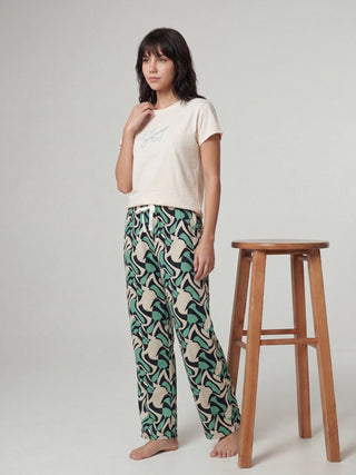 A woman in beige pajamas with a green and cream wavy print and white slippers stands with her hands in her pockets.