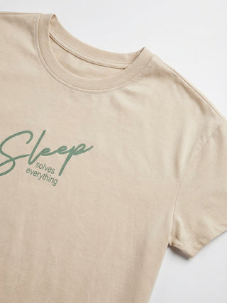 A woman in a beige t-shirt with a green and cream wavy print saying "Sleep solves everything" stands with her hands in her pockets.