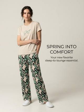 A woman in beige pajamas with a green and cream wavy print and white slippers stands with her hands in her pockets.