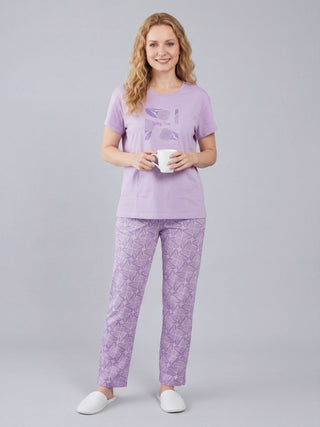 A woman stands holding a mug wearing a lavender t-shirt with a tulip graphic and matching lavender patterned pants.