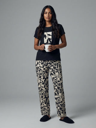 A woman stands holding a mug, wearing a black t-shirt with a leaf print and beige pants with a black leaf print.