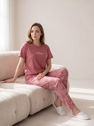 A woman wearing pink pajama pants with a floral print and a pink short-sleeved top with "Bloom" embroidered on it, sitting on a cream sofa.