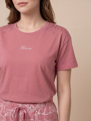 A woman stands, wearing a pink t-shirt with a white "Bloom" text and floral accents on the shoulders.
