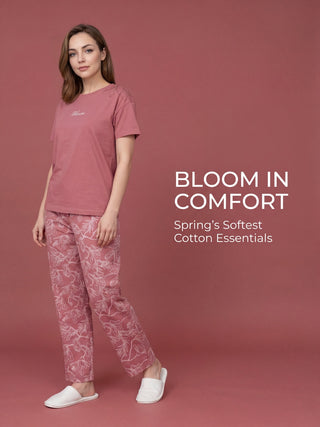 A woman stands, wearing a pink t-shirt with white "Bloom" text and floral accents on the shoulders, and matching pink floral patterned pants.