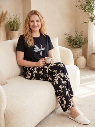A woman wearing a black t-shirt with a floral print and matching black pants with a floral print, sitting on a couch with a mug.