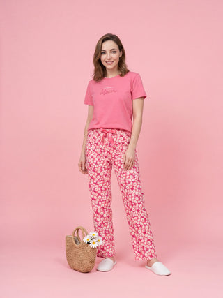 Woman in pink t-shirt with "bloom" graphic and pink floral print pajama pants, standing with a straw bag and white slippers.