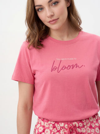 A woman in a pink t-shirt with "You were made to bloom" printed on it and floral patterned shorts is shown from the chest down, posed in a studio setting.