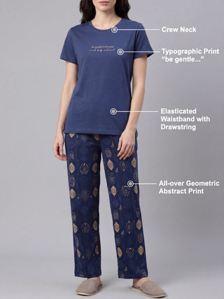 Woman wearing a navy blue crew neck pajama set with a typographic print on the top and an all-over geometric abstract print on the pants, and slippers.