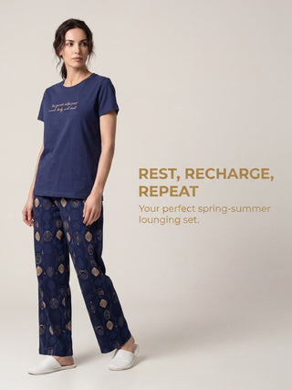 A woman models a navy blue pajama set with a gold-colored "Be gentle with your mind, body and soul" print on the t-shirt and a gold geometric pattern on the pants, while looking to the side with white slippers on.