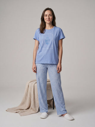 A woman stands posing in a blue t-shirt with white "La Dolce Vita" text and blue patterned pajama pants.