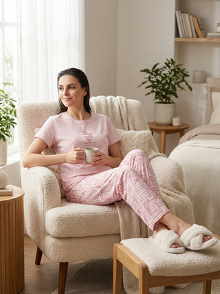 A woman is lounging in a plush armchair, wearing a pink pajama set with a geometric print and holding a mug.