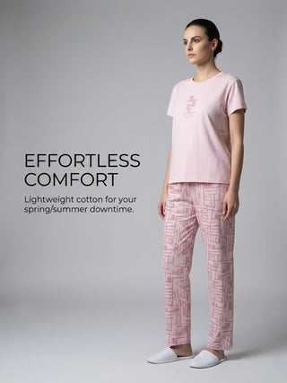 A woman stands looking right, wearing pink printed pajamas and white slippers.