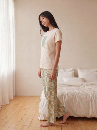 Woman in a light beige t-shirt with abstract print and green and beige checkered pajama bottoms, standing in profile.