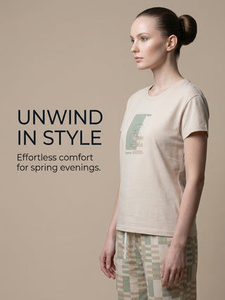 Woman in a light beige t-shirt with abstract print and green and beige checkered pajama bottoms, standing in profile.