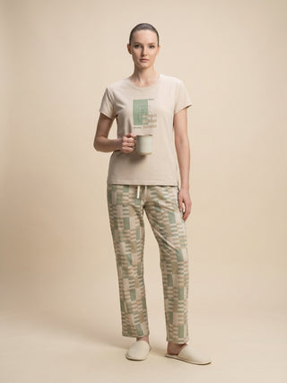 Woman in a beige t-shirt with a graphic print and green and beige checkered pajama pants, holding a mug.