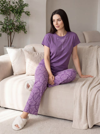 A woman in a purple printed pajama set with white fuzzy slippers sits on a cream couch, with her legs crossed.
