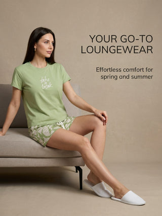 A woman poses on a couch wearing a light green t-shirt with "take a break" printed on it and matching checkered shorts.