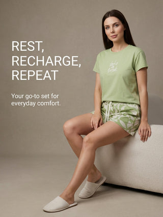A woman in a light green t-shirt with "take a break" graphic and matching green geometric shorts with white accents, sits on a white textured bench wearing cream slippers.