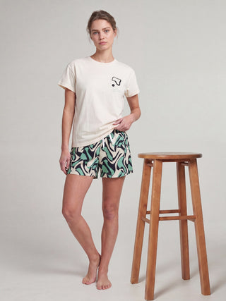 A woman wearing a light beige t-shirt with a black graphic and multi-colored geometric shorts, stands with her legs crossed and wears white slippers.