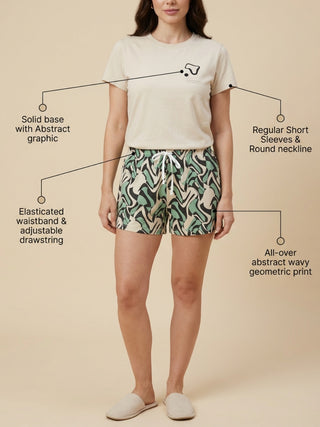 A woman wearing a light beige t-shirt with a black graphic and multi-colored geometric shorts, stands with her legs crossed and wears white slippers.
