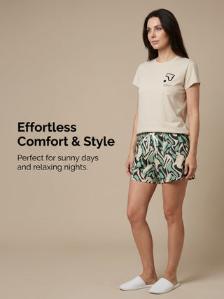 A woman in a light beige t-shirt with a black graphic print and mint green and black abstract print shorts poses with her left leg forward.