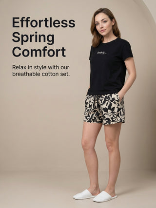 Woman in a black t-shirt with "Simplicity" printed in white and matching black and white floral print pajama shorts, standing with hands in pockets and wearing white slippers.
